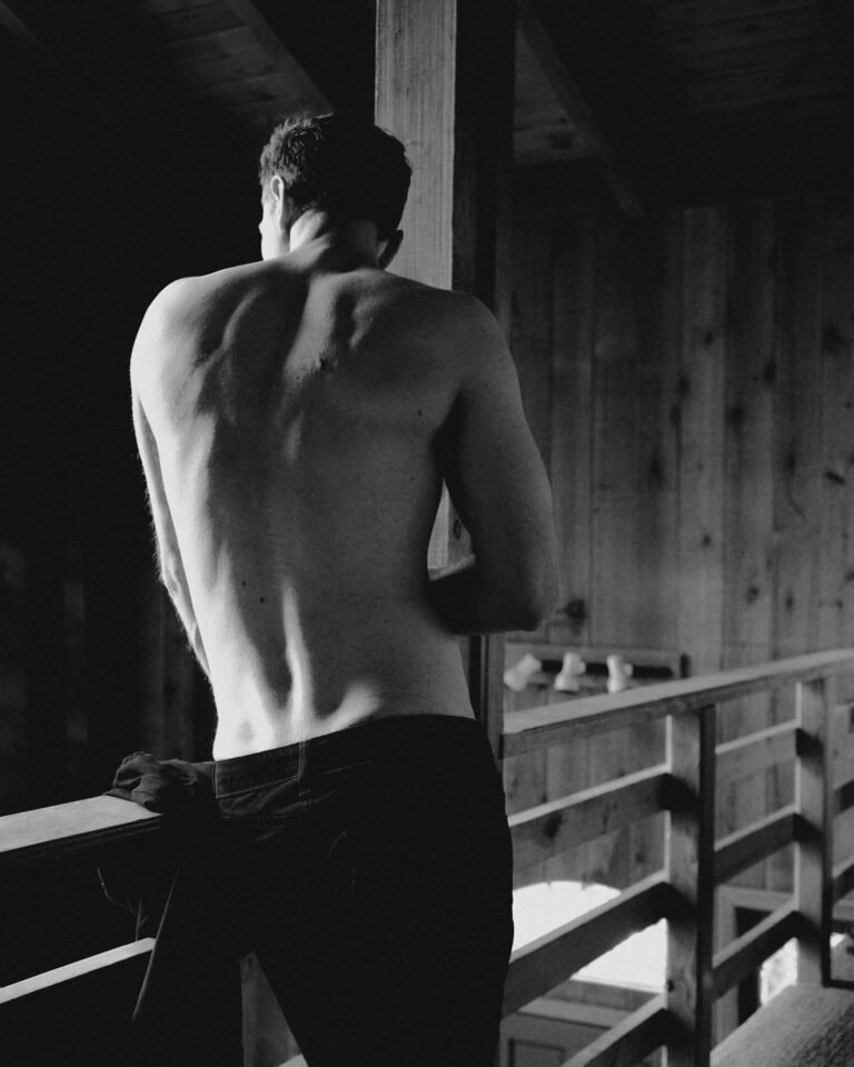 Shirtless man seen from behind, leaning on a wooden balcony railing